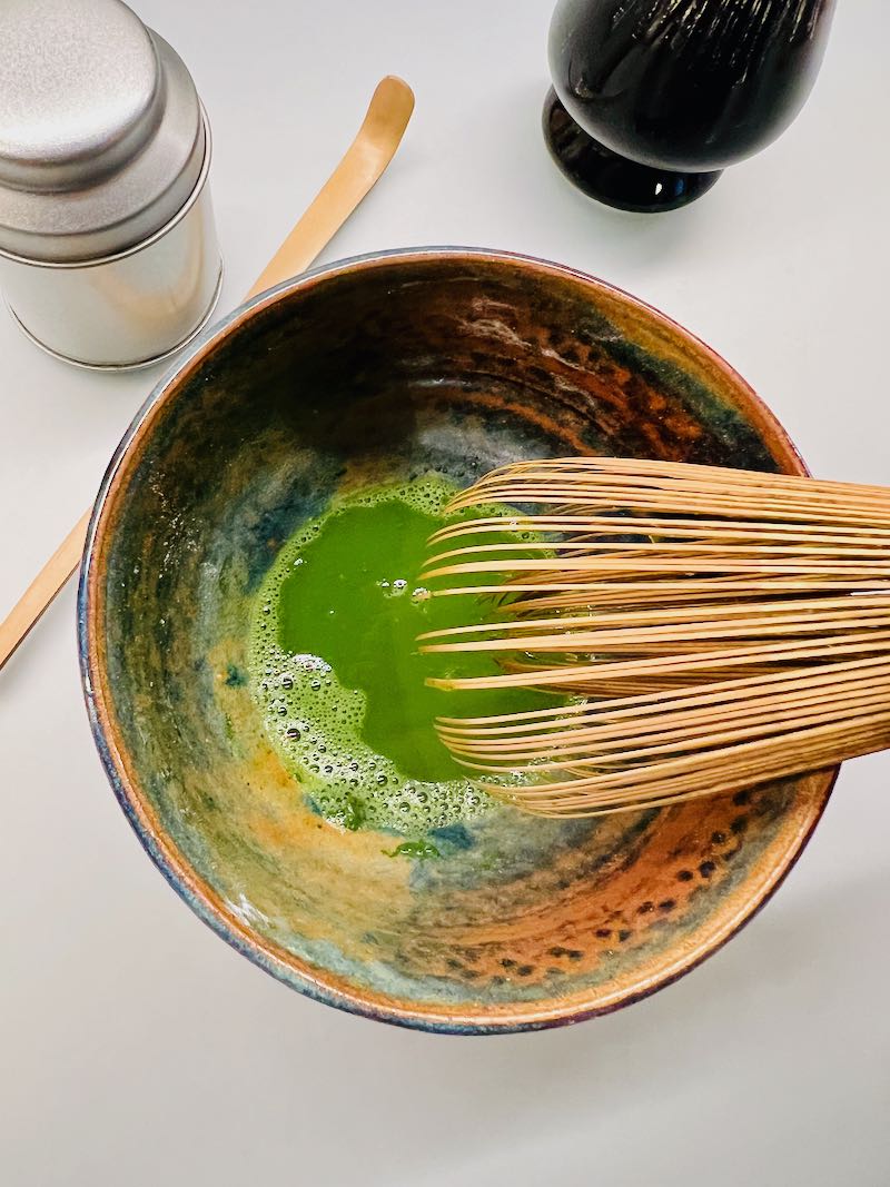 Met een matcha klopper worden de aanwezige klontjes glad gestreken. Boven de kom liggen een matcha bamboelepel en theeblikje.