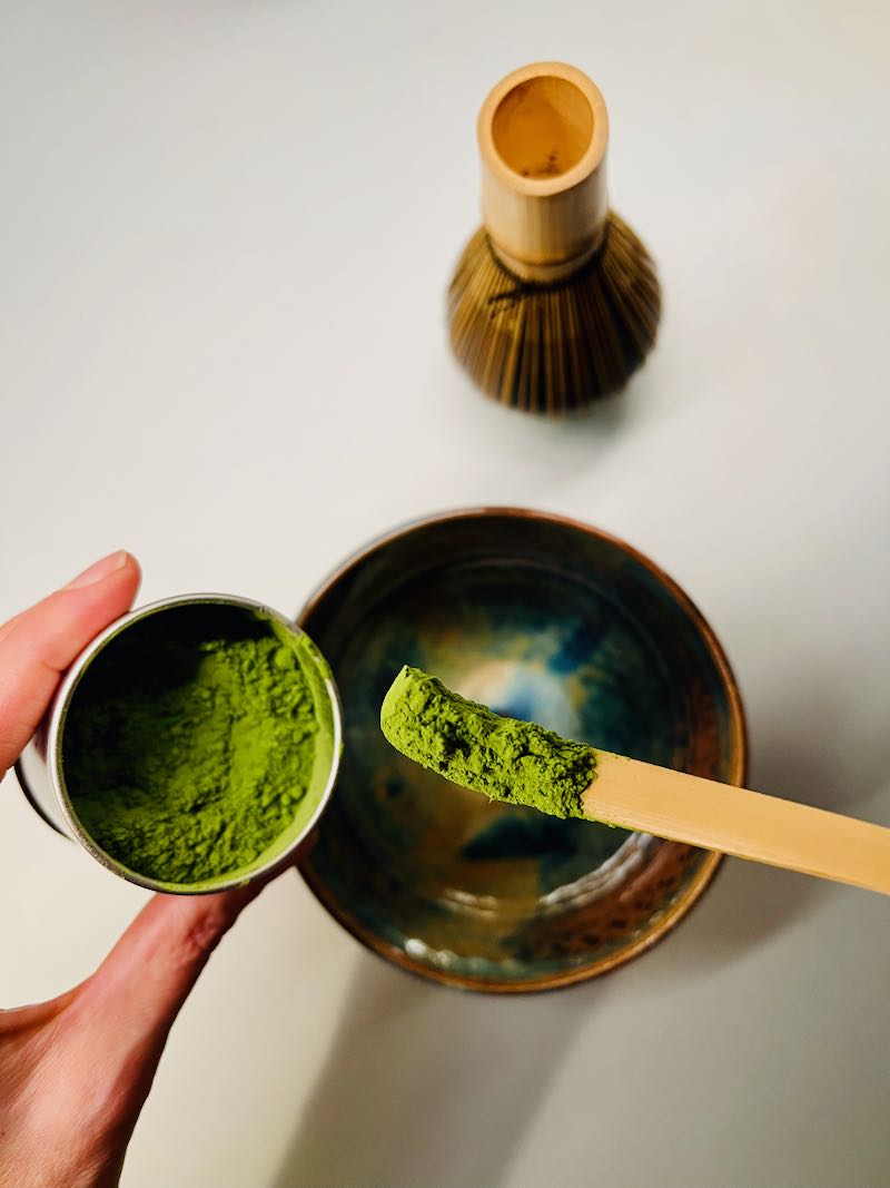 A small amount of matcha tea powder is placed in a bowl with a bamboo teaspoon. Above is a matcha brush.