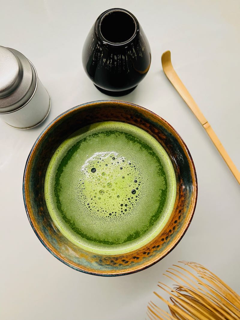 Een mok Matcha, met een rechtsboven een matcha borstel houder en matcha lepel. Rechtsonder een matcha borstel.