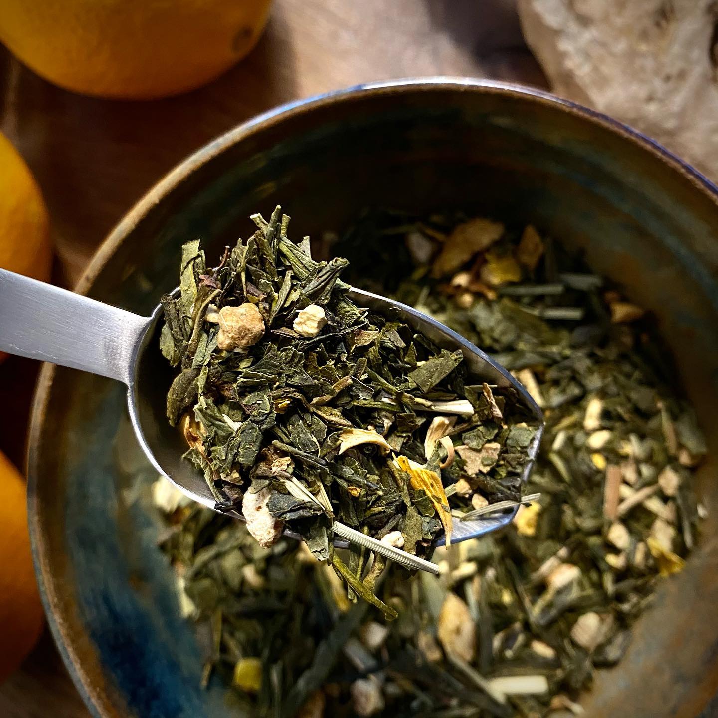 A spoon full of Sencha green tea leaves, enriched with orange slices and ginger, above a ceramic bowl, with a blurred background of an orange and wooden surface.