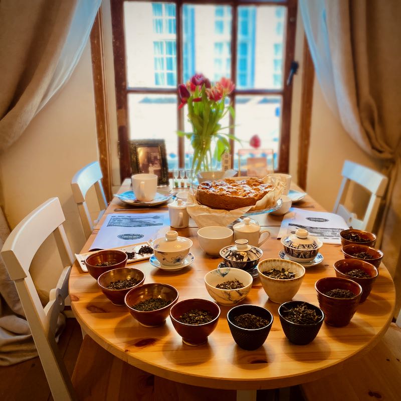 A cozy tea tasting setup with a variety of tea mugs and saucers on a wooden round table, a freshly baked cake in the center, teapots, and flowers in a vase by the window overlooking the street